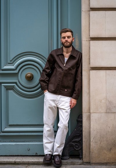 Chaqueta marrón con cremallera, cuello y bolsillos delanteros, combinada con pantalones blancos y zapatos marrones, frente a un fondo de puerta azul.