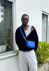 Μαύρο καρντάγκαν με άνετη εφαρμογή πάνω από γκρι κοντομάνικο, συνδυασμένο με γκρι φόρμες και μια μικρή μπλε τσάντα ώμου.