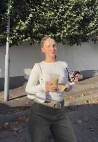 Hvit langermet topp, høy midje mørke jeans, klar kopp med drikke, lys håndveske, og svarte solbriller i hånden, med bladete bakgrunn.