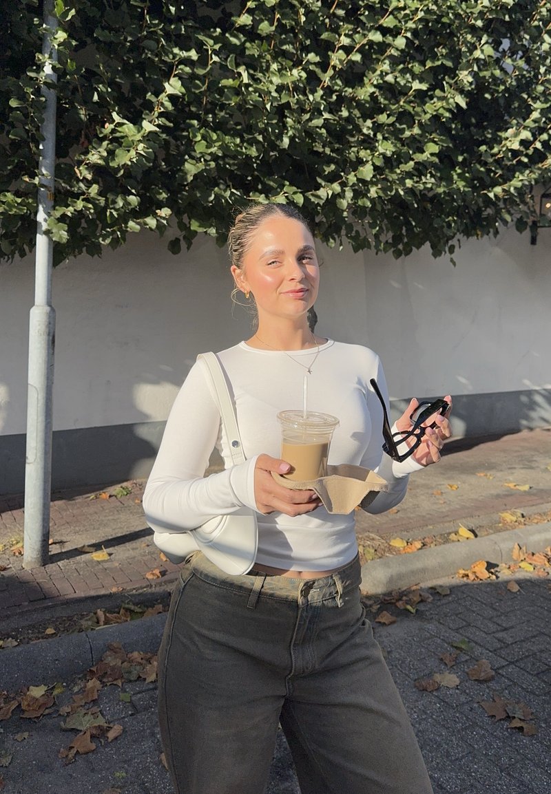 Weißes Langarmoberteil, hochgeschnittene dunkle Jeans, klarer Becher mit Getränk, leichte Handtasche und schwarze Sonnenbrille in der Hand, mit blättrigem Hintergrund.