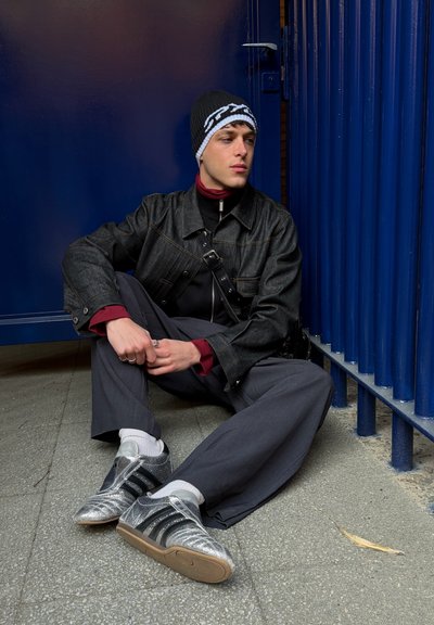 Joven con chaqueta de mezclilla oscura, pantalones grises, zapatillas plateadas y gorro de punto sentado en pavimento gris cerca de una valla metálica azul y una puerta, mirando de lado.