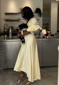 Un vestito fluido di raso giallo con morbide maniche a campana, abbinato a tacchi a piattaforma argentati. Un cane peloso bianco e nero tenuto tra le braccia.