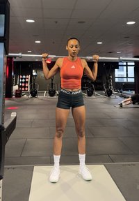 Atleta a levantar uma barra de pesos em interior. Ela usa uma blusa laranja sem mangas, calções pretos com o texto "PRO NIKE" e sapatilhas brancas com detalhes verdes.