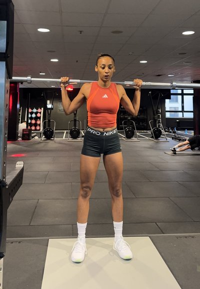 Atleta levantando una barra en interiores. Lleva una camiseta sin mangas naranja, pantalones cortos negros con el texto "PRO NIKE" y zapatillas blancas con detalles en verde.