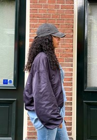 Zwarte puffy bomberjack, denim overalls, blauwe spijkerbroek en een grijze pet. Krullend haar valt voorbij de schouders. Bakstenen muur op de achtergrond.