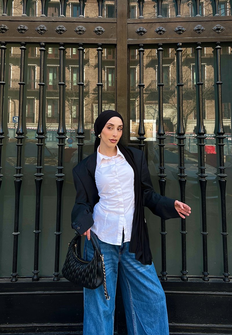 Vrouw met een zwarte hoofddoek en blazer, wit overhemd, ruime blauwe spijkerbroek, die een zwarte gewatteerde tas vasthoudt, staand tegen een zwart ijzeren hek.