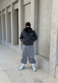 Chaqueta impermeable negra, pantalones grises holgados, zapatillas deportivas plateadas y un pasamontañas negro. De pie contra una pared de hormigón texturizada.