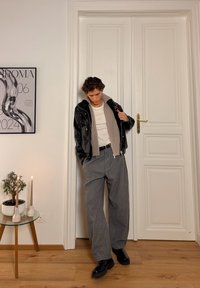 Veste en cuir noire avec col côtelé gris, t-shirt blanc, pantalon gris à jambes larges et chaussures noires. En arrière-plan, des bougies et une plante.