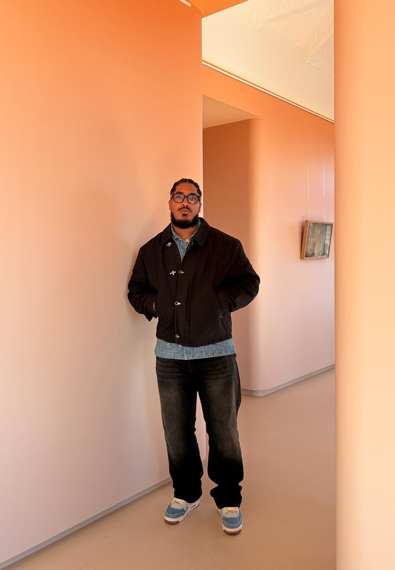Homme avec lunettes et cheveux tressés porte une veste noire, une chemise bleue, un jean et des baskets, debout dans un espace intérieur moderne aux tons orange.