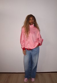 Jaqueta desportiva rosa com fecho éclair e detalhes em vermelho, com três riscas brancas nas mangas, combinada com calças de ganga largas azuis claras e ténis rosas.