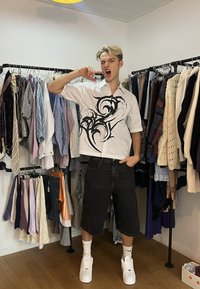 Jeune homme en shorts noirs et chemise blanche à motifs posant avec la main près du visage à l'intérieur d'un magasin de vêtements avec des portants de vêtements.