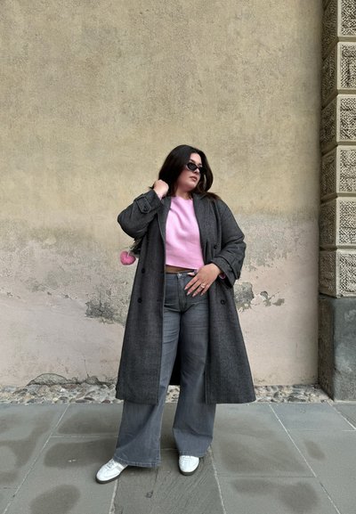 Abrigo negro de espiga largo sobre un suéter corto rosa claro, jeans acampanados grises y zapatillas blancas. Gafas de sol negras y un charm de pompón.