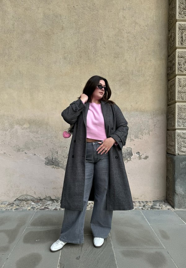 Langer schwarzer Fischgrätmantel über einem hellrosa Cropped-Sweater, grauen Schlagjeans und weißen Sneakers. Schwarze Sonnenbrille und ein Pom-Pom-Anhänger.