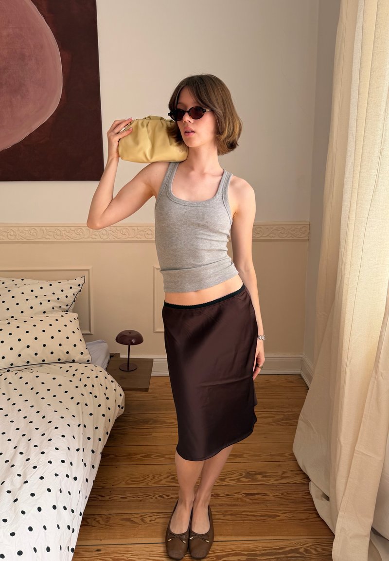 Jeune personne portant des lunettes de soleil, un débardeur gris et une jupe foncée tient un petit sac jaune près du visage, debout dans une chambre avec une literie à pois.