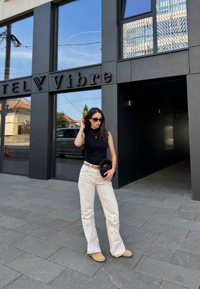 Top sin mangas negro, pantalones de pierna ancha color blanco roto, zapatos beige, sosteniendo un bolso negro. Fachada de un edificio moderno al fondo con grandes ventanas.