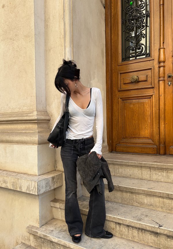Une femme vêtue d’un haut blanc et d’un jean foncé se tient sur des marches en pierre, près d’une porte en bois ouvragée, avec une veste et un sac en bandoulière.