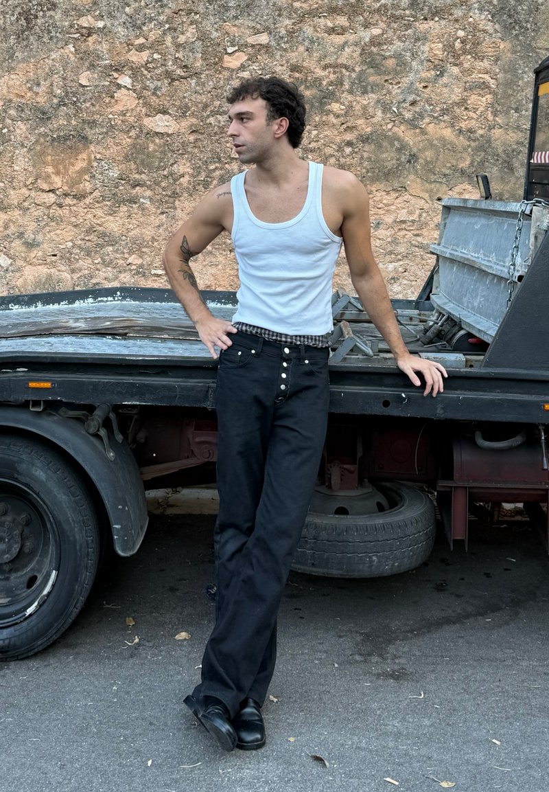 Top bianco senza maniche, cintura a scacchi, pantaloni neri con bottoni e scarpe nere. Appoggiato a un camion con uno sfondo testurizzato.