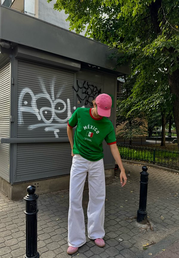 Personne portant un t-shirt vert Mexique, un pantalon blanc, une casquette et des chaussures roses, debout sur un trottoir près d'un bâtiment couvert de graffitis et d'arbres.