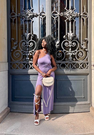 Vestido maxi morado con escote asimétrico, abertura lateral y detalles de cordones. Bolso blanco con correa de cadena, sandalias de plataforma con acentos de cinta.