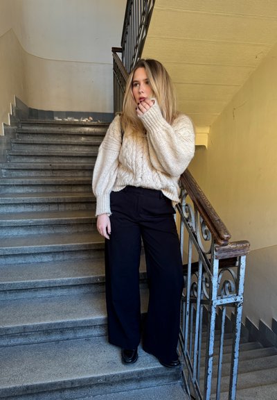 Joven mujer con suéter beige y pantalones negros de pie en una escalera junto a una barandilla de metal, mirando hacia un lado con la mano cerca de la boca.