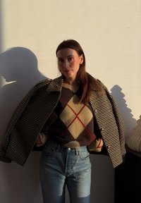 Femme aux longs cheveux bruns portant une veste à carreaux par-dessus un pull à losanges et un jean bleu, debout contre un mur beige avec des ombres.