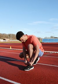 Sapatilhas atléticas pretas com detalhes brancos, uma t-shirt justa laranja e calções azuis claros; pessoa a amarrar os atacadores numa pista de corrida vermelha.