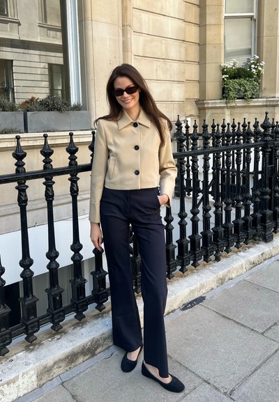 Chaqueta corta beige con botones negros, llevada sobre pantalones oscuros y zapatos de ballet negros, de pie junto a una barandilla de hierro forjado negro.