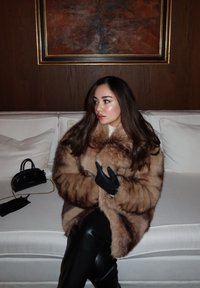 Cappotto marrone in pelliccia oversize con dettagli scuri, abbinato a pantaloni e guanti in pelle nera. Piccola borsa nera accanto su divano bianco.