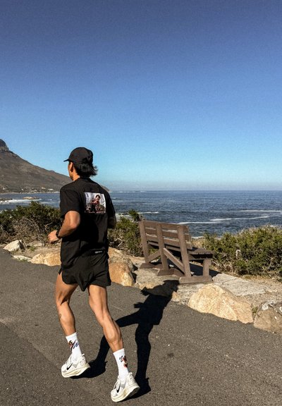 Hombre corriendo despacio por un sendero costero cerca de un banco con vistas al océano y las montañas bajo un cielo azul claro.