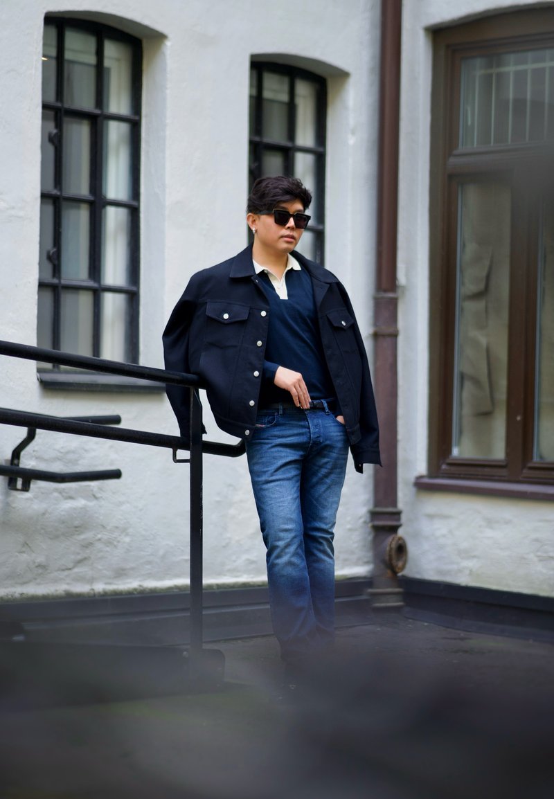 Giacca di jeans nera con bottoni argentati, maglione blu navy e colletto chiaro, abbinati a jeans blu; occhiali da sole completano il look.