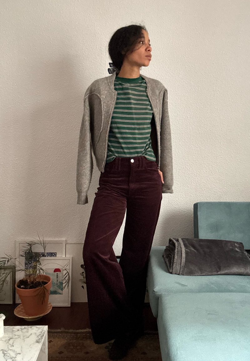 Chaqueta bomber gris, camiseta de rayas verdes y pantalones de pana burdeos de pierna ancha. El modelo está de pie frente a una pared blanca texturizada con muebles.