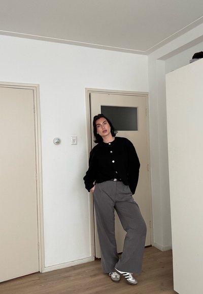 Persona con cabello oscuro se apoya en la puerta de una habitación vistiendo un suéter negro abotonado, pantalones grises de pierna ancha y zapatillas blancas con rayas negras.