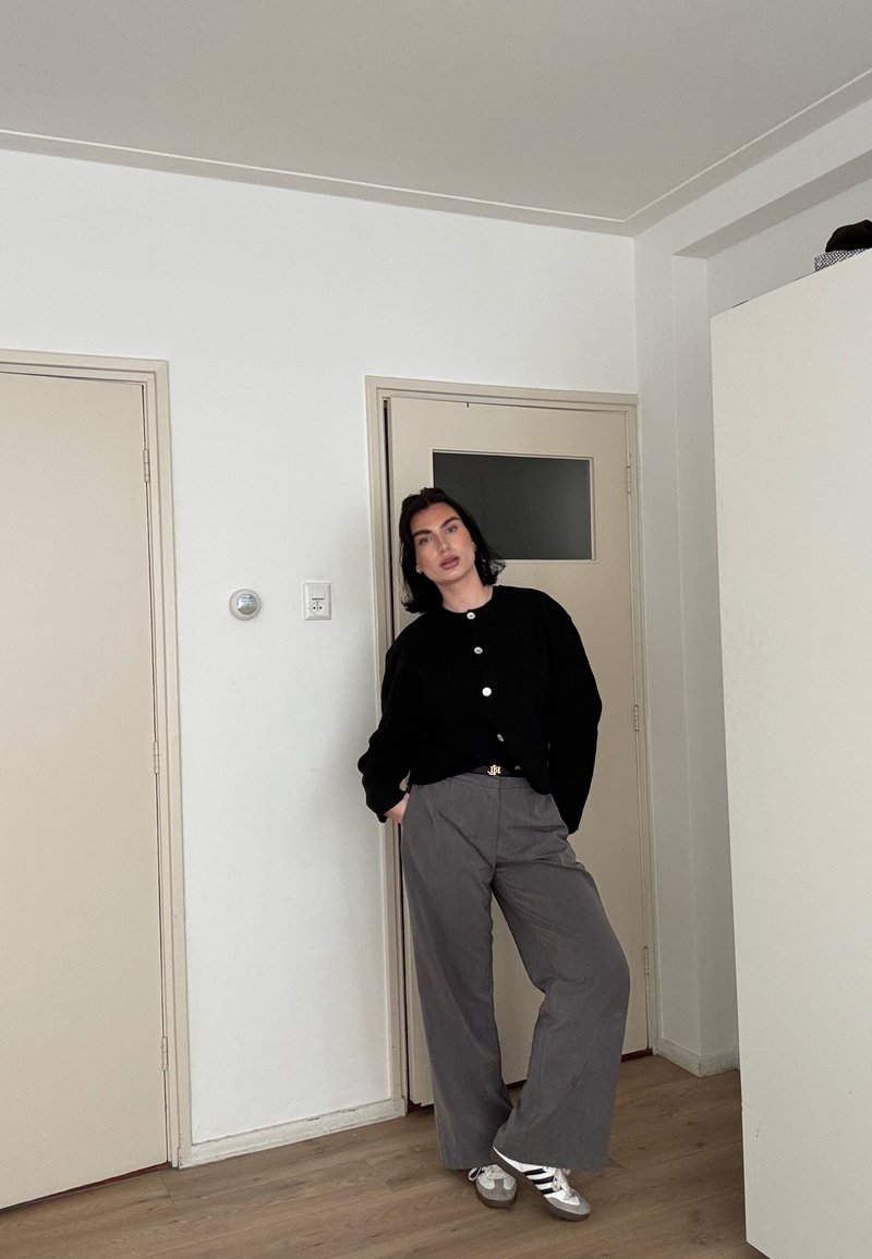 Personne aux cheveux foncés appuyée contre la porte dans une pièce, portant un pull noir boutonné, un pantalon large gris et des baskets blanches avec des rayures noires.