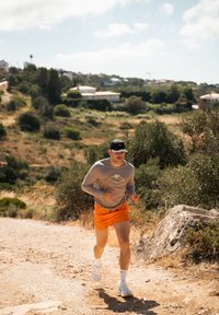 Atletický muž v šedém tričku s dlouhým rukávem s grafickým motivem hory a oranžových šortkách běží po prašné cestě mezi zelení pod jasnou oblohou.