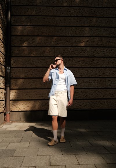 Camisa de manga corta azul claro con una camiseta blanca, pantalones cortos de color crema y zapatos de lona beige. De pie sobre una superficie pavimentada contra una pared texturizada.