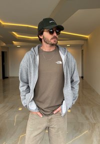 Grå zip-up hoodie over en brun t-shirt, med tan bukser. Tilbehør inkluderer en grøn corduroy-kasket og runde solbriller. Indendørs setting.