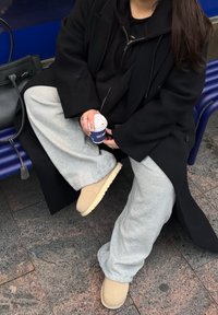 Sort oversized frakke, sort hættetrøje, grå joggingbukser, beige slip-on støvler. Holder en lille blå kaffekop med teksten "CAFE".