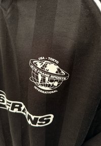 Schwarzes Sporttrikot mit vertikalen Streifen. Es zeigt eine weiße Globusgrafik mit dem Text "USA - TOKYO" und "IETS FRANS SPORTS INTERNATIONAL."