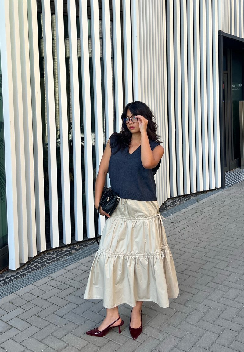 Top sem mangas azul-marinho combinado com uma saia em camadas de cetim creme, acessorizado com uma bolsa preta e sapatos de salto vermelhos, situado contra um fundo listrado.