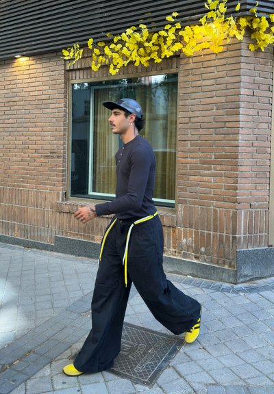 Pantalones anchos negros con cordones amarillos, combinados con una camiseta negra de manga larga y zapatos amarillos, llevados por una persona que camina al aire libre.