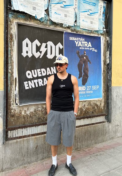 Camiseta de tirantes negra, pantalones cortos grises plisados, calcetines blancos y mocasines negros. Hombre de pie frente a una pared texturizada con carteles de conciertos.