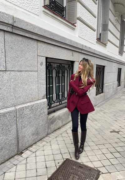 Blazer marrón con hombros estructurados, top negro y pantalones negros ajustados. Botas marrones hasta la rodilla. Pared de piedra gris texturizada de fondo.