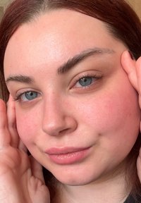 Nærbilde av en persons ansikt med middels hudtone, blå øyne og mørkerødt hår. Naturlig sminke, jevn hud med en svak rosafarge.