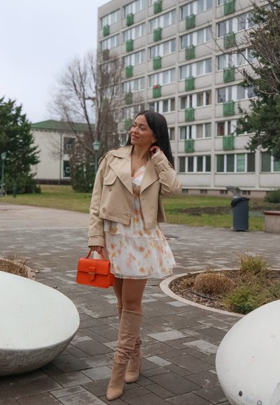 Mujer con chaqueta beige, vestido floral y botas hasta la rodilla caminando por un camino pavimentado, sosteniendo un bolso naranja, con edificios residenciales de fondo.