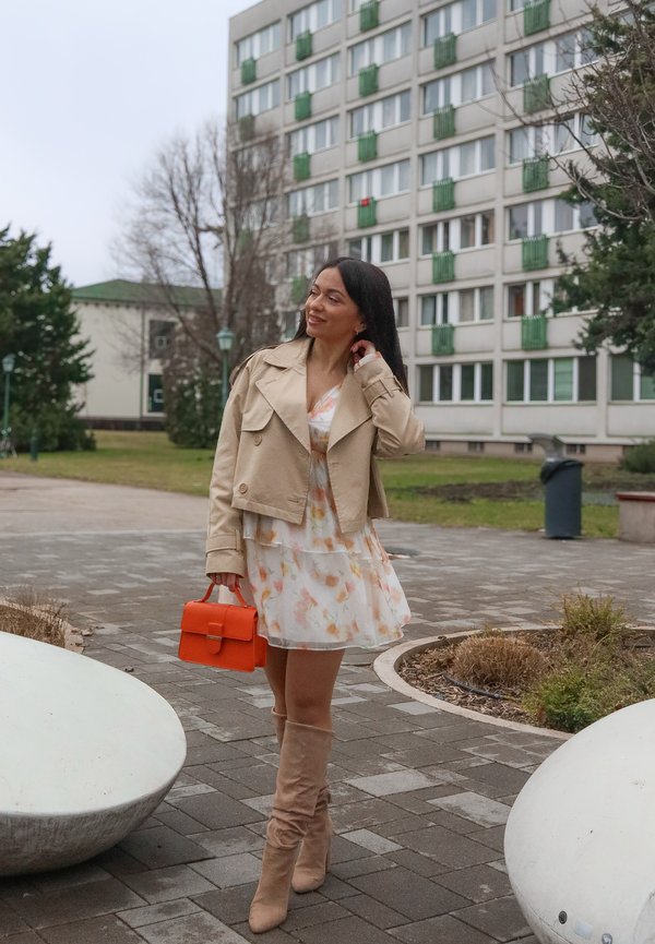 Femme en veste beige, robe fleurie, bottes hauteur genoux marchant sur un chemin pavé, tenant un sac à main orange, avec des immeubles résidentiels en arrière-plan.