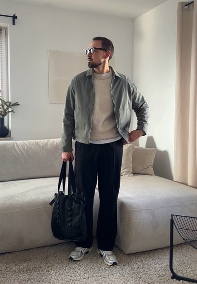 Hombre con gafas, chaqueta gris, suéter beige, pantalones negros, zapatillas blancas, sosteniendo un bolso tote negro, de pie en una sala de estar luminosa.