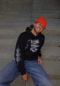 Melns džemperis ar kapuci ar grafisko printu priekšpusē un baltām zvaigžņu sprādzienu dizainiem uz piedurknēm. Kombinēts ar gaiši zilām džinsa biksēm un oranžu cepuri.