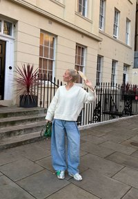 Pull blanc oversize en maille avec un col en V, jean denim bleu clair ample, baskets argentées, et un sac à main vert. Plantes et architecture visibles.