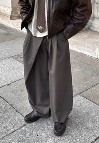 Bruine leren jas over een licht shirt en een bruin gestreepte stropdas; brede grijze broek; donkere schoenen; staand op een stenen trottoir.