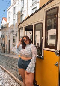 Top corto de encaje floral azul con mangas acampanadas, combinado con pantalones cortos de mezclilla de tiro alto. El fondo presenta un tranvía amarillo vintage.